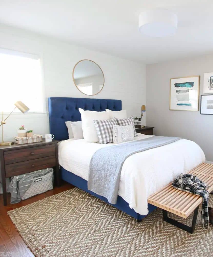 Blue bed frame, dark brown bedside table, and light wood foot rest with very light gray walls